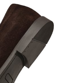Mocasín de ante marrón oscuro con suela de goma negra y punta del tacón texturizada, inclinado para mostrar detalles laterales y de la suela.