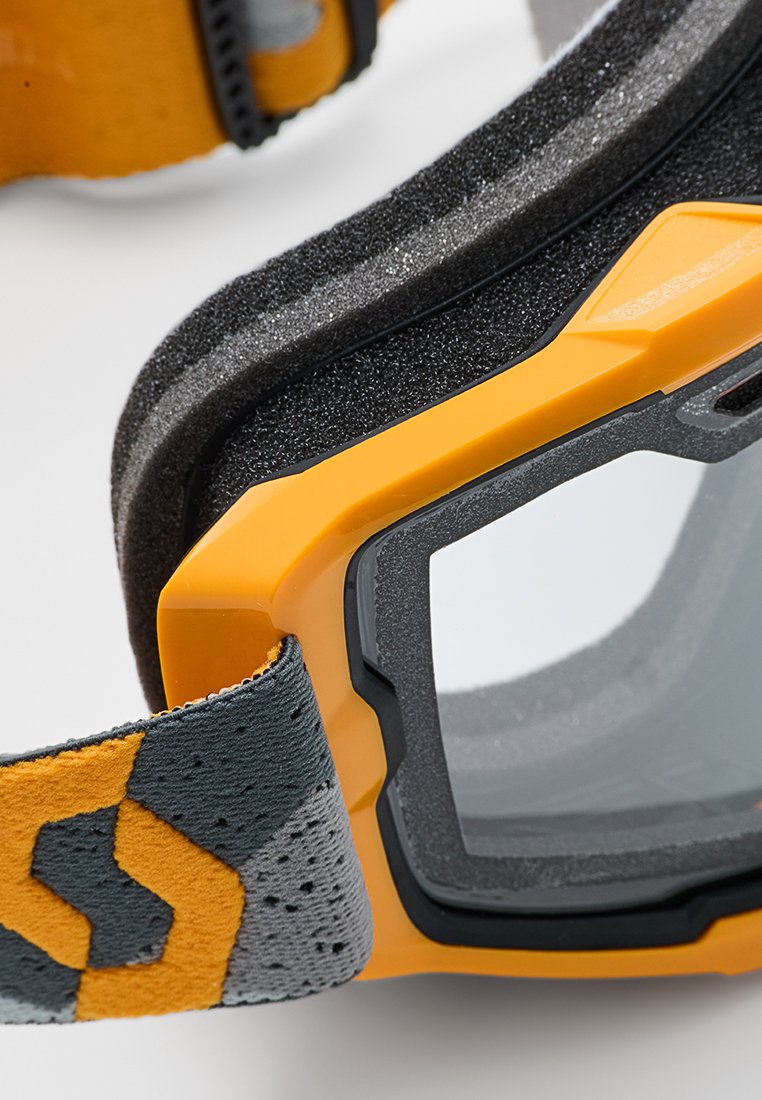 Gafas de protección con un marco amarillo y gris, acolchado de espuma negra, lente transparente y correa ajustable con un patrón texturizado.