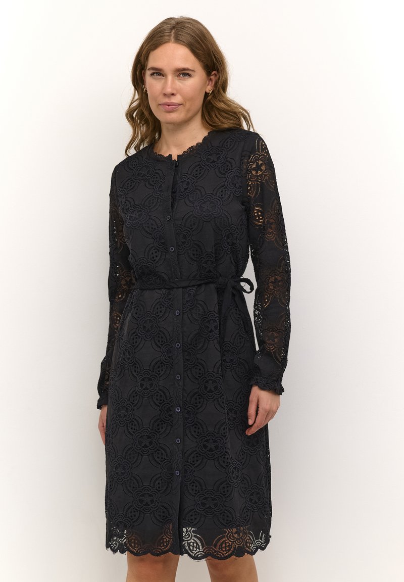 Svart spetsklänning med långa ärmar, knappar framtill och ett justerbart knyte i midjan. Har blommönster och en kant med vågformat avslut.