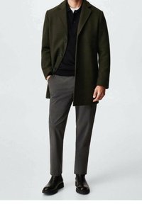 Donkergroen overjas met knopen, gecombineerd met een zwarte poloshirt en grijze maatwerkbroek; afgemaakt met zwarte enkellaarsjes.