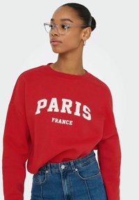 Rode sweatshirt met witte tekst "PARIS" en "FRANCE", ronde halslijn, lange mouwen, gecombineerd met high-waisted blauwe denim jeans.