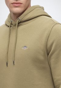 Homme portant un sweat à capuche beige avec cordons et un petit patch logo GANT sur la poitrine, recadré sous le nez.
