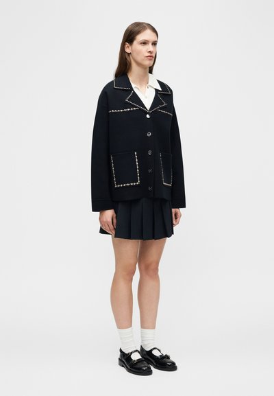 Γυναίκα ντυμένη με μαύρο τζάκετ με τρουκς πάνω από ένα λευκό πουκάμισο με γιακά, μαύρη πλισέ φούστα, λευκές κάλτσες και μαύρα παπούτσια Mary Jane, στέκεται όρθια.