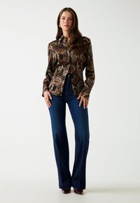 Donna in blusa scura a paisley, jeans a vita alta blu scuro a zampa di elefante e décolleté borgogna a punta, su sfondo bianco semplice.