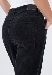 Zwarte corduroybroek met een ribbeltextuur, voorzien van achterzakken en een label op de tailleband. Slim fit met subtiele stikseldetails.