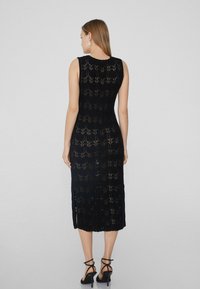 Schwarzes Häkelkleid mit ärmellosem Design, intricatem Muster, knieumspielender Länge und seitlichen Schlitzen. Getragen mit schwarzen Sandalen mit Riemen.