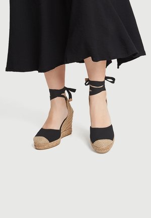 Sandalias de cuña de lona negra con suela de cuerda de yute, con punta redondeada y lazos en el tobillo hechos de tela negra.