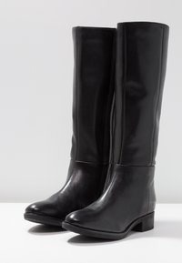 Bottes hautes en cuir noir avec une texture lisse, un bout arrondi, un petit talon bloc et des éléments de design minimalistes.