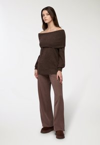 Brązowy sweter typu off-the-shoulder z prążkowanym dekoltem, zestawiony z jasnobrązowymi, wzorzystymi spodniami szerokimi i brązowymi butami wsuwanymi.
