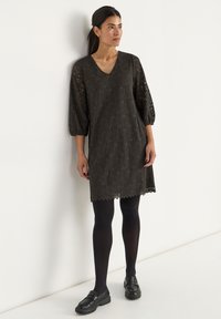 Spitzenkleid in Dunkelbraun mit V-Ausschnitt, dreiviertellangen Puffärmeln, gewelltem Saum, getragen über schwarzen Strumpfhosen und kombiniert mit schwarzen Slippern.