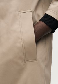 Cappotto impermeabile beige con texture liscia, tasca rettangolare e polsini neri a coste. Primo piano della manica e dettaglio laterale.