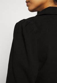 Chaqueta de mezclilla negra con un cuello estructurado, detalle de hombros abullonados y textura acanalada, que presenta un acabado suave.