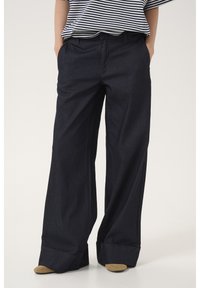 Pantaloni in denim scuro a gamba larga con una texture liscia, dotati di piega frontale, tasche laterali e orli arrotolati. Indossati con una maglietta a righe.