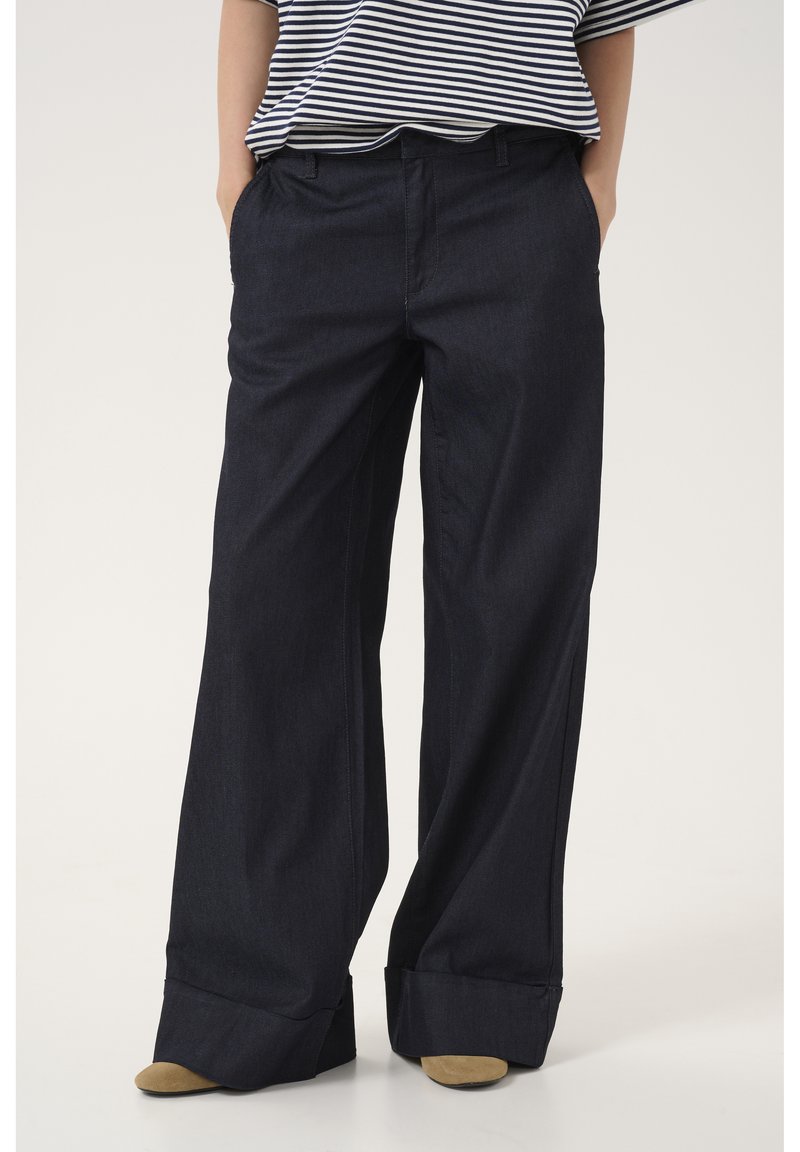Pantaloni in denim scuro a gamba larga con una texture liscia, dotati di piega frontale, tasche laterali e orli arrotolati. Indossati con una maglietta a righe.