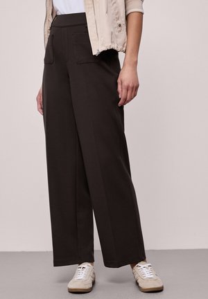 Personne portant un pantalon noir taille haute, jambes larges avec poches avant, associé à des baskets blanches et une veste légère beige.