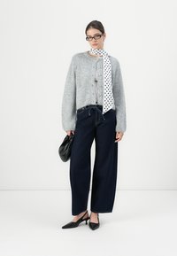 Lichtgrijze fuzzy cardigan met knopen, donkerblauwe wijde jeans, een witte sjaal met stippen, een zwarte handtas en zwarte puntige hakken.