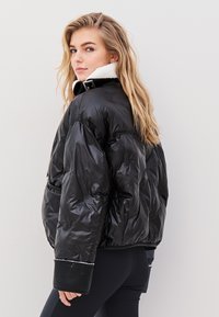 Fekete puffkabát fényes felülettel, fehér gyapjú gallérral, bordázott mandzsettákkal és oldalsó zsebekkel. Könnyű és jól megformált kialakítás.