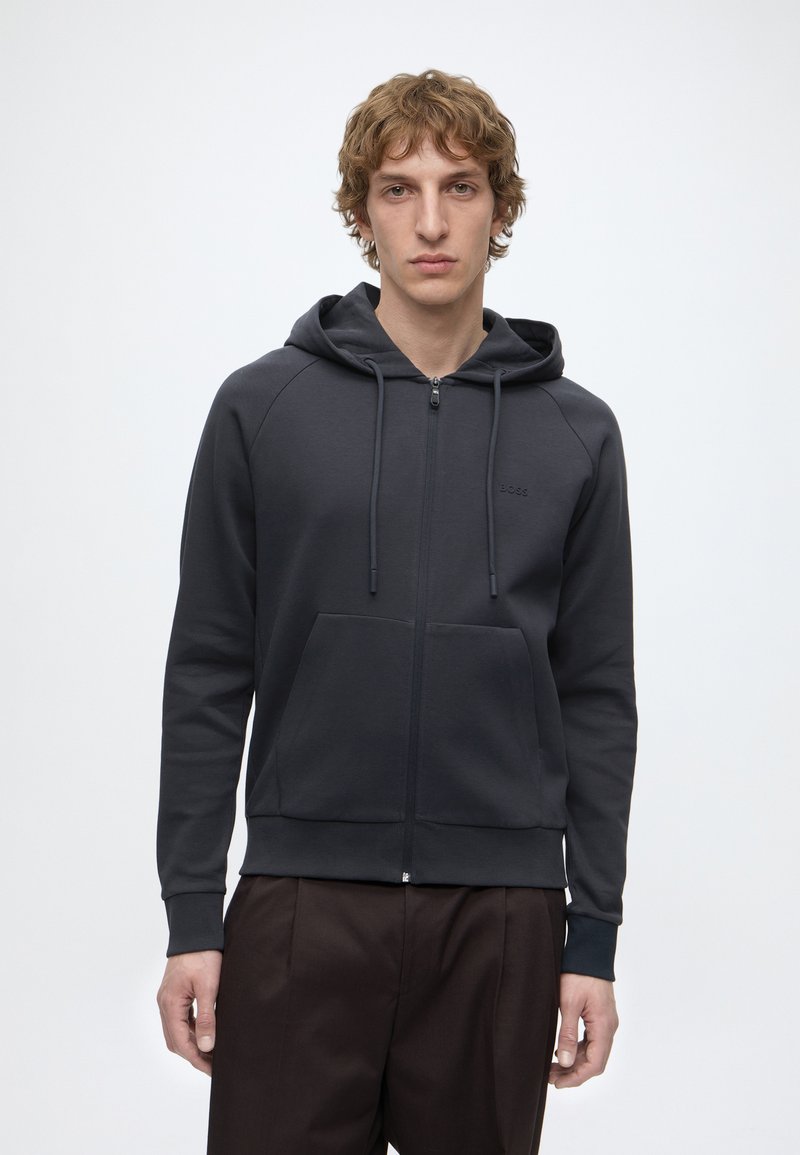 Jeune homme aux cheveux bouclés châtain clair portant un sweat à capuche noir zippé avec des poches avant et un pantalon foncé, debout devant un fond blanc uni.