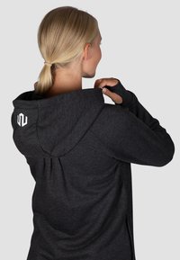 Schwarze Kapuzenjacke mit weicher Textur, rundem Kapuzenkragen, Daumenlöchern in den Ärmeln und einer Faltenoptik auf der Rückseite. Logo auf der Kapuze sichtbar.