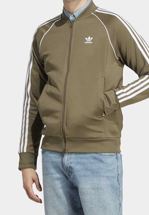 Mies pukeutunut oliivinvihreään Adidas-vetoketjutakkiin valkoisilla raidoilla hihoissa ja vaaleansiniset farkut, käsi takin taskussa.