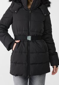 Black puffer jacket with zip closure, belted waist, side pockets, and a detachable faux fur hood. Smooth texture and quilted design.