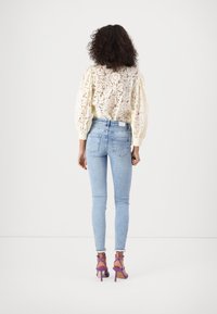 Blusa de encaje en crema claro con mangas abullonadas, combinada con jeans ajustados azul claro y sandalias de tacón moradas. Vista trasera centrada en los detalles.