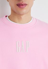 Světle růžová mikina vyrobená z bavlněné směsi; má bílé vyšívané logo "GAP" na hrudi. Ribovaný kulatý výstřih, měkká textura.