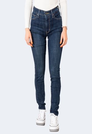 Jean slim bleu foncé porté avec des baskets blanches à plateforme et un haut blanc à manches longues, montrant de la taille aux pieds sur un fond clair.