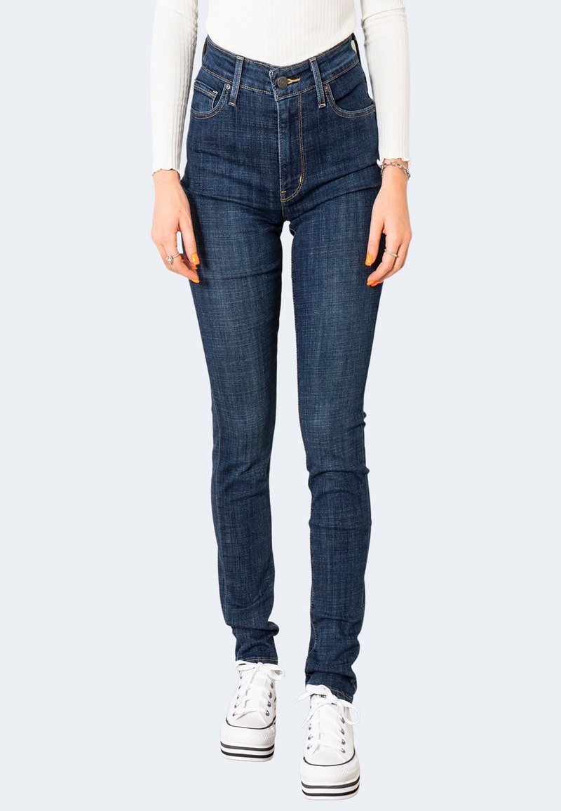 Jean slim bleu foncé porté avec des baskets blanches à plateforme et un haut blanc à manches longues, montrant de la taille aux pieds sur un fond clair.