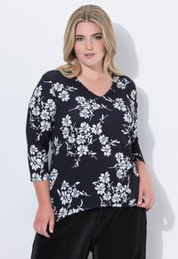 Frau mit blonden Haaren, die ein schwarzes Oberteil mit weißem Blumenmuster und schwarze Hose trägt, steht vor einem weißen Hintergrund.