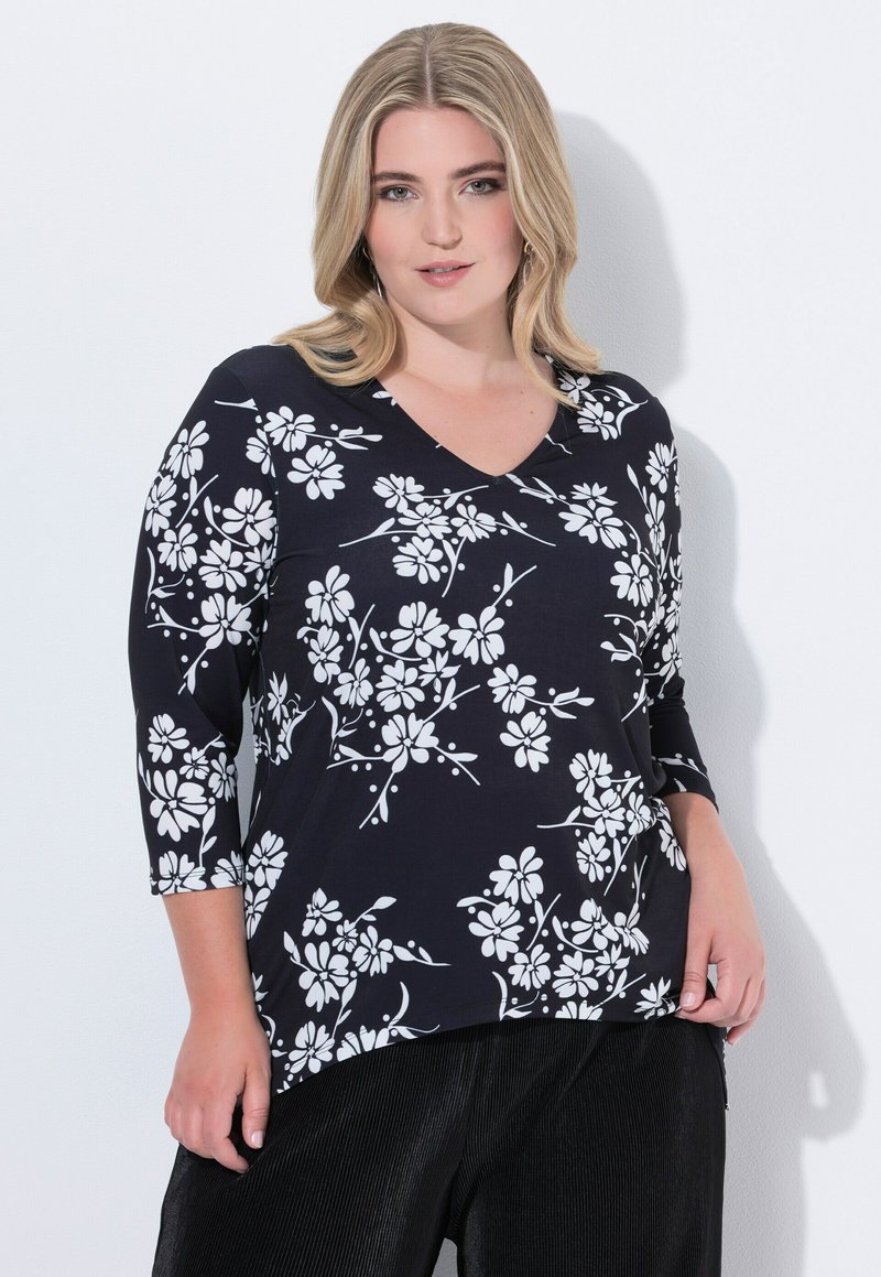 Frau mit blonden Haaren, die ein schwarzes Oberteil mit weißem Blumenmuster und schwarze Hose trägt, steht vor einem weißen Hintergrund.