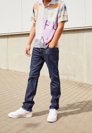 Mężczyzna w koszuli z krótkim rękawem w abstrakcyjne kolory, ciemnoniebieskich dżinsach i białych trampkach stojący na brukowanym zewnętrznym chodniku.