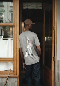 Hombre con camiseta gris con un dibujo de una persona y un perro en la parte trasera, de pie en una puerta con marco de madera y paneles de vidrio.