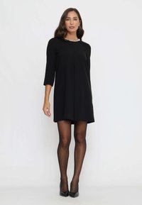 Schwarzes A-Linien-Kleid mit Dreiviertelärmeln, das eine gefältelte Vorderseite und einen silbernen Akzent am Ausschnitt aufweist, kombiniert mit transparenten schwarzen Strumpfhosen.