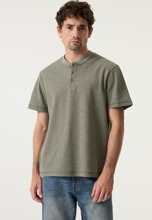 Maglietta henley verde oliva a maniche corte con tessuto strutturato, caratterizzata da una pattina con tre bottoni e una vestibilità comoda. Indossata con jeans azzurri.