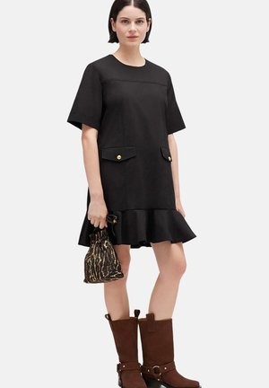 Donna che indossa un vestito nero a maniche corte con tasche frontali e orlo con balze, che tiene in mano una piccola borsa decorata, abbinato a stivali marroni fino a metà polpaccio.