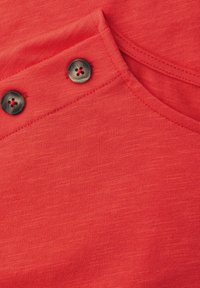 Czerwona tkanina z dwoma okrągłymi ciemnymi guzikami przyszytymi ukośnie w pobliżu przeszytego narożnika szwu.