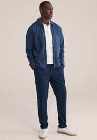 Marineblauw jack met twee borstzakken, gecombineerd met marineblauwe broek en witte polo met structuur. Schoeisel bestaat uit witte sneakers met beige zolen.