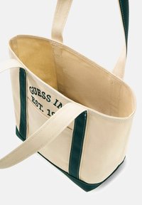Borsa a tracolla in tela di colore crema con accenti verde scuro. Presenta un logo stampato e testo. Interno aperto con fodera morbida e spallacci robusti.