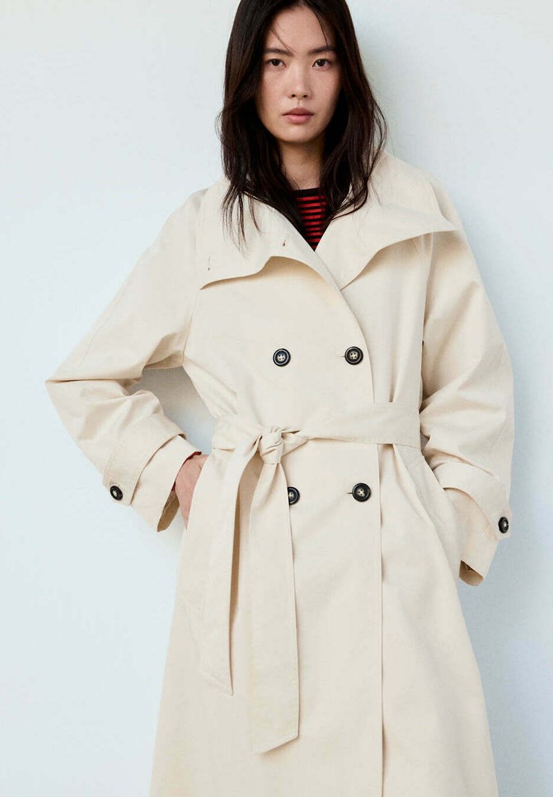 Jeune femme aux longs cheveux foncés portant un trench-coat beige ceinturé avec des boutons noirs et les mains dans les poches, sur un fond uni.