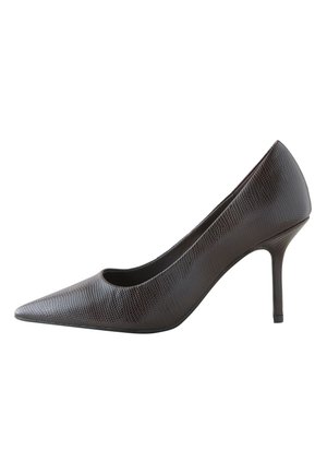 Donna con capelli ricci che indossa un maglione bianco con bottoni e pantaloni corti marroni, mettendo scarpe nere con tacco alto davanti a una parete liscia.