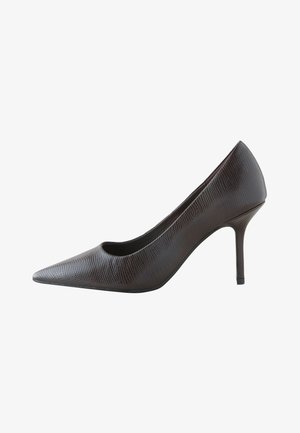 Donna con capelli ricci che indossa un maglione bianco con bottoni e pantaloni corti marroni, mettendo scarpe nere con tacco alto davanti a una parete liscia.