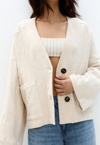 Vrouw in een open beige knoopjasje over een wit gestreept crop top en blauwe jeans, met hand die het jasje gesloten houdt.