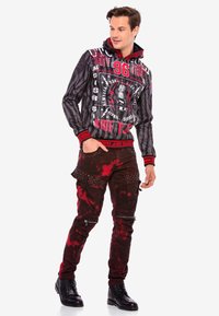 Schwarze und graue gestreifte Kapuzenjacke mit roten Akzenten und grafischen Prints, kombiniert mit dunkelroten Jeans, die Farbkleckse, Nieten und Reißverschlüsse aufweisen. Schwarze Stiefel.