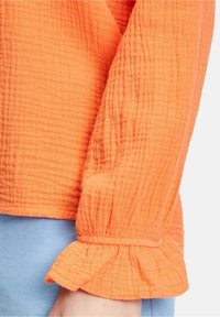 Blusa arancione brillante realizzata in tessuto texturizzato e increspato. Presenta un polsino a ruches e un orlo dritto, abbinata a un tessuto azzurro chiaro sottostante.