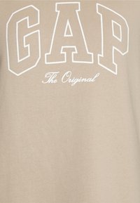 Beżowa bluza z dużym, białym napisem "GAP" i mniejszym, kursywą napisem "The Original". Miękka, gładka tekstura z luźnym krojem.