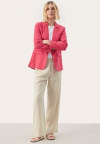 Różowa lniana marynarka z dwoma przednimi kieszeniami i złotymi guzikami, w połączeniu z białym t-shirtem i beżowymi spodniami typu wide-leg, uzupełniona sandałami.