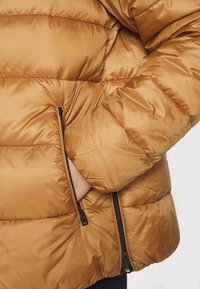 Personne portant une doudoune marron avec une main partiellement dans une poche zippée sur le côté, sur un fond uni.
