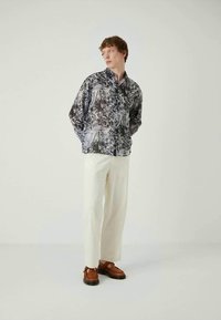 Πουκάμισο με κουμπιά και ασπρόμαυρο αφηρημένο σχέδιο, σε συνδυασμό με λευκό παντελόνι και καφέ παπούτσια, τοποθετημένο σε φωτεινό φόντο.