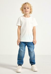 Hvid bomuldst-shirt, blå denimjeans, elastiske manchetter og hvide sneakers med velcrolukning. Barn står oprejst og ser fremad.